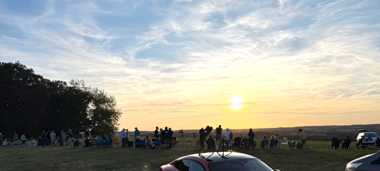 Crowd with telescopes and camp chairs on a hilltop meadow at sunset during the August 2026 solar eclipse